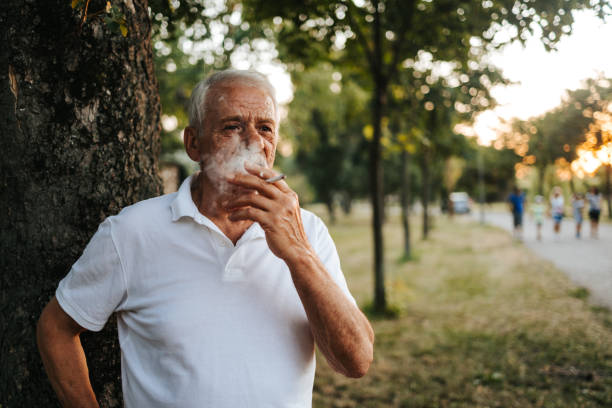 Senior man standing under the tree in public park at sunset. Enjoys cigarettes