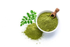 Overhead view of Matcha tea powder in a white bowl isolated on white background. Some Matcha powder is spilled on the background and Moringa leaves are beside the bowl. A wooden serving scoop complete the composition. High resolution 42Mp studio digital capture taken with Sony A7rII and Sony FE 90mm f2.8 macro G OSS lens