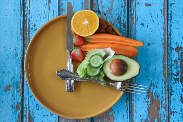 Stock photo showing an intermittent fasting concept depicted by a plate and cutlery forming a clock face and hands with the smallest section on the plate being filled with a pile of almonds, half an orange, strawberry cut in two, a sliced carrot, lettuce leaves, slices of cucumber and half an avocado displaying it's large seed.