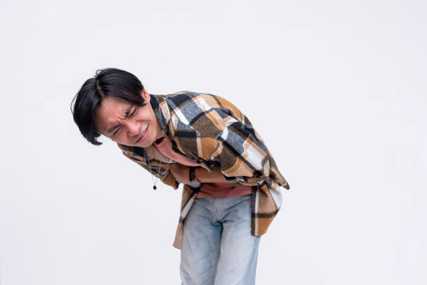 A young asian man winces in extreme pain, clutching his stomach. Cramped and hurting. Isolated on a white background.